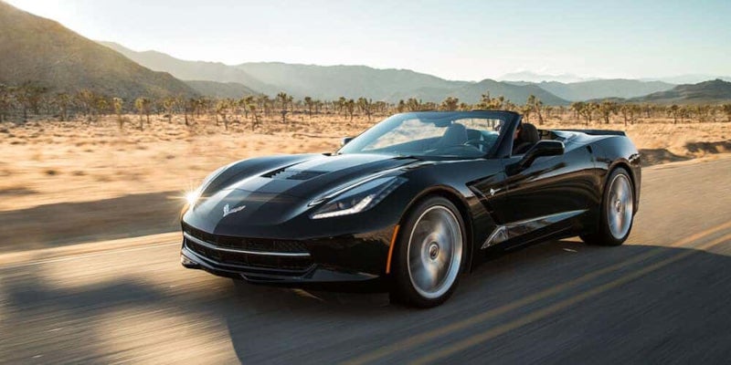Black convertible sports car driving on a desert road with mountains in the background.
