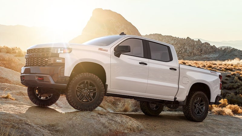 White Chevrolet Silverado pickup truck on rocky off-road terrain with mountains and sun in the background.