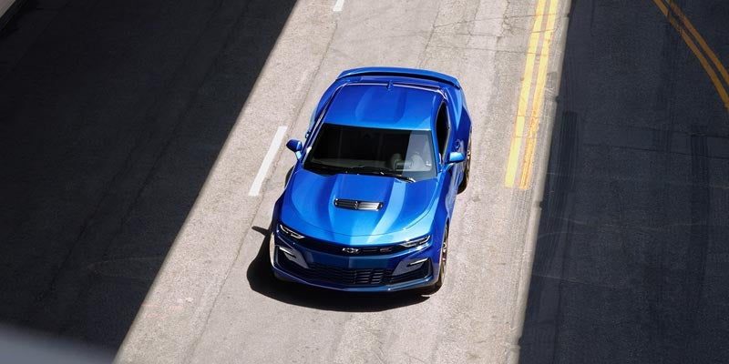 Overhead view of a blue Chevrolet Camaro on an asphalt road with strong sunlight and shadows.