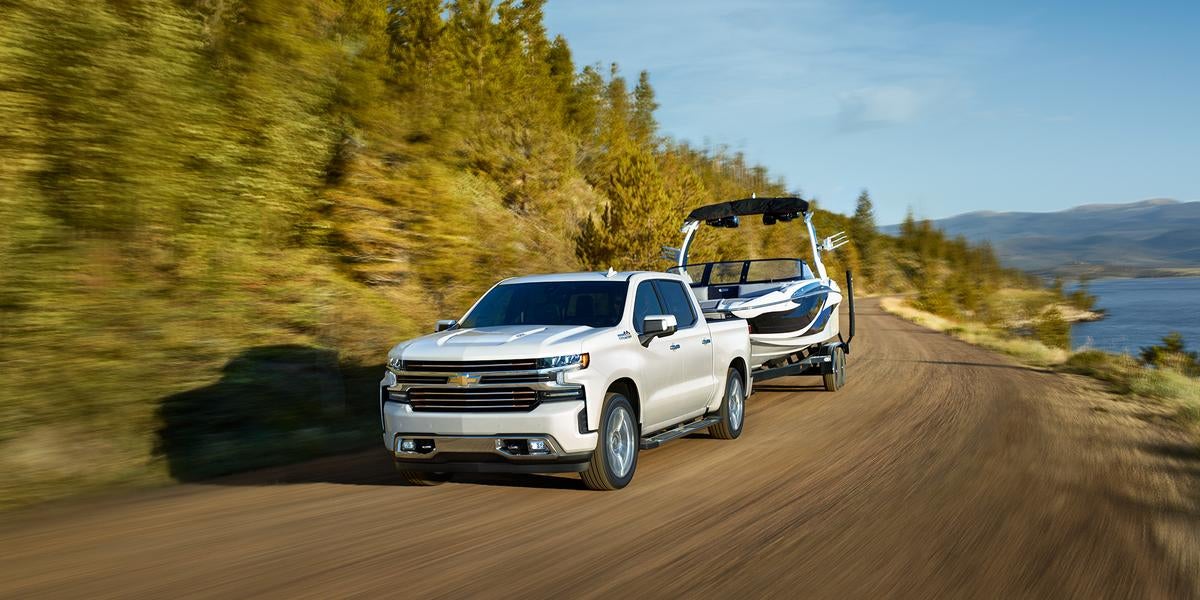 White pickup truck towing a boat on a trailer down a dirt road next to a lake with green trees.