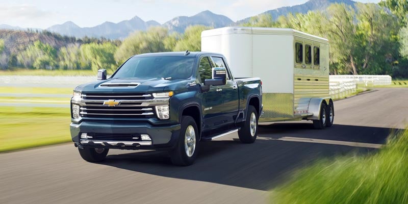 Dark blue Chevy Silverado truck towing a white and silver horse trailer on a rural road with mountains in the background.
