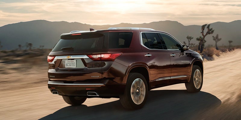 Maroon Chevrolet Traverse SUV driving on a dirt road in a desert landscape with mountains and Joshua trees.