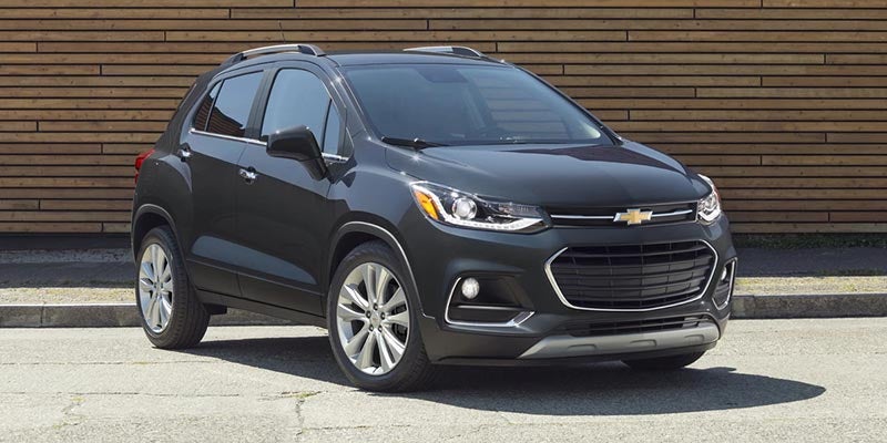 Dark gray Chevrolet Trax compact SUV parked on asphalt in front of a wooden slatted wall, angled slightly right.