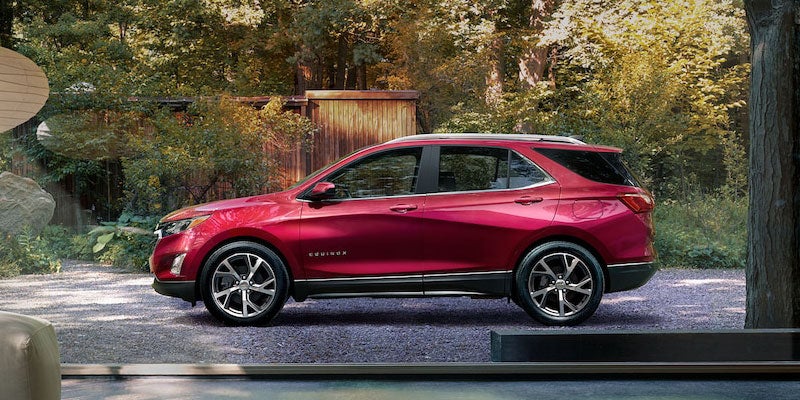 Red Chevy Equinox SUV parked on a gravel driveway amidst trees, viewed through a large window from a modern interior.