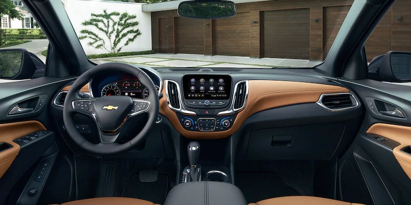View from inside a Chevrolet car showing the tan and black dashboard, steering wheel, infotainment system, and a house outside.