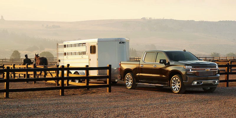 Brown pickup truck towing a white horse trailer at a ranch, with a person and horse in the background under hazy light.