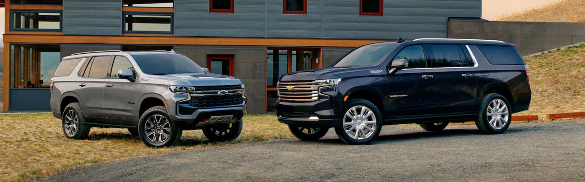 Two Chevrolet SUVs, a silver Tahoe Z71 and a dark blue Suburban High Country, parked in front of a modern home.