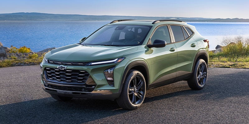 Green Chevrolet Trax SUV parked on asphalt with a large blue lake, rocky shore, and hills in the background.