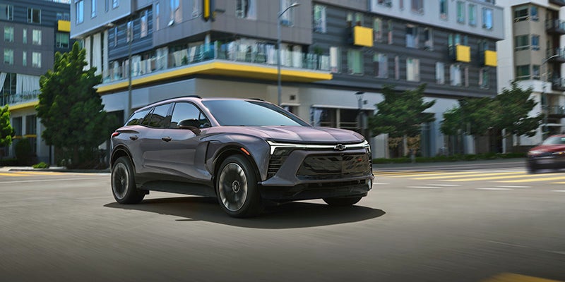 A dark gray Chevrolet Blazer EV drives on a city street with modern buildings in the background.
