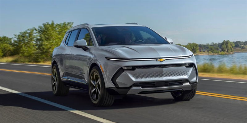 Light gray Chevrolet Equinox EV SUV driving on a road with a lake and trees in the background.