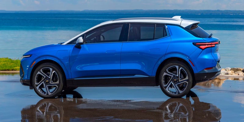 Blue and white electric SUV parked sideways near a body of water, reflecting on the wet ground.