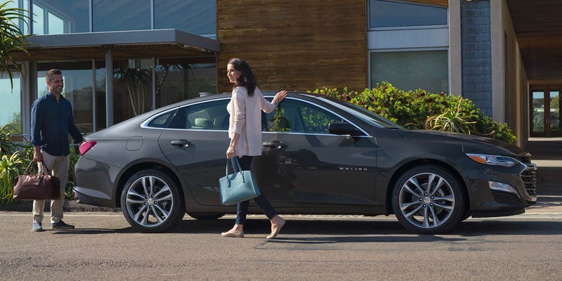 A man and woman with luggage stand next to a dark gray Chevrolet Malibu sedan parked in front of a modern building.