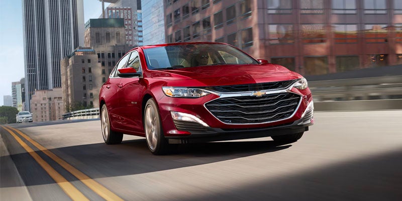 Red Chevrolet Malibu sedan driving on a multi-lane city road with blurred buildings in the background.