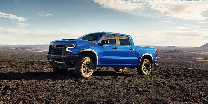Blue Chevrolet Silverado pickup truck parked on dark rocky terrain, overlooking a vast forest landscape under a cloudy sky.