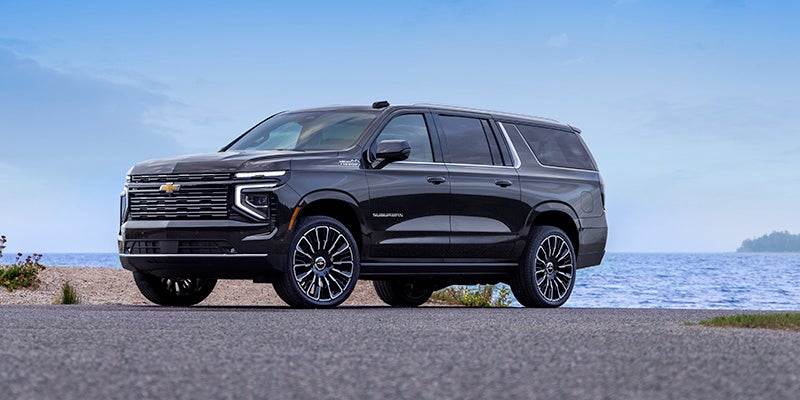 Black Chevrolet Suburban SUV parked on an asphalt road next to a large body of water under a blue sky.