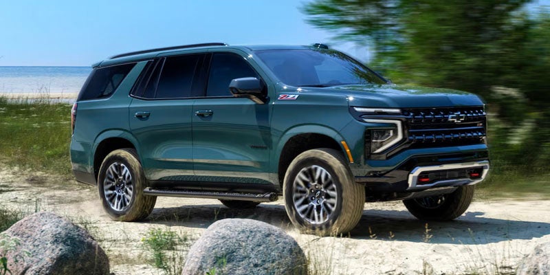 Dark green Chevy Tahoe Z71 SUV driving on a dirt path with rocks, water, and grass in the background.