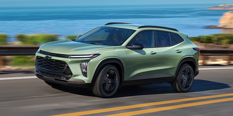 Sage green SUV driving on a coastal road with a blue ocean and distant land in the background.