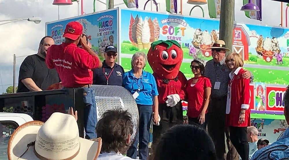 A group of people and a cheerful strawberry mascot pose for a photo in front of an ice cream stand.