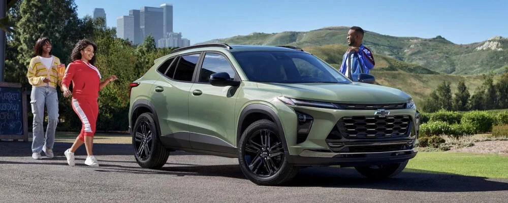 Light green Chevrolet Trax SUV parked on a road with three people around it, city buildings and mountains in the background.