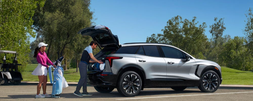 A man loads golf clubs into the open trunk of a silver SUV, while a woman holds another golf bag nearby on a golf course.