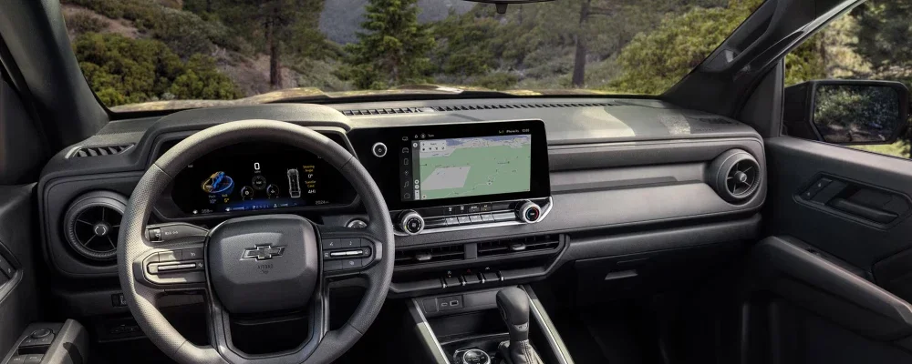 The modern car interior features a steering wheel, digital gauge cluster, and large touchscreen with a map, looking out to trees.