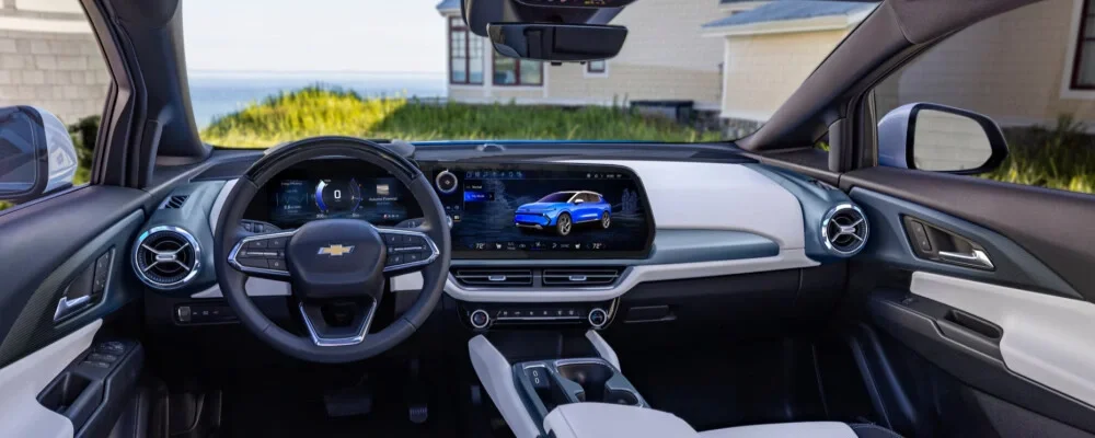 Modern car interior with steering wheel, digital gauges, and large central touchscreen displaying a blue SUV, ocean view outside.
